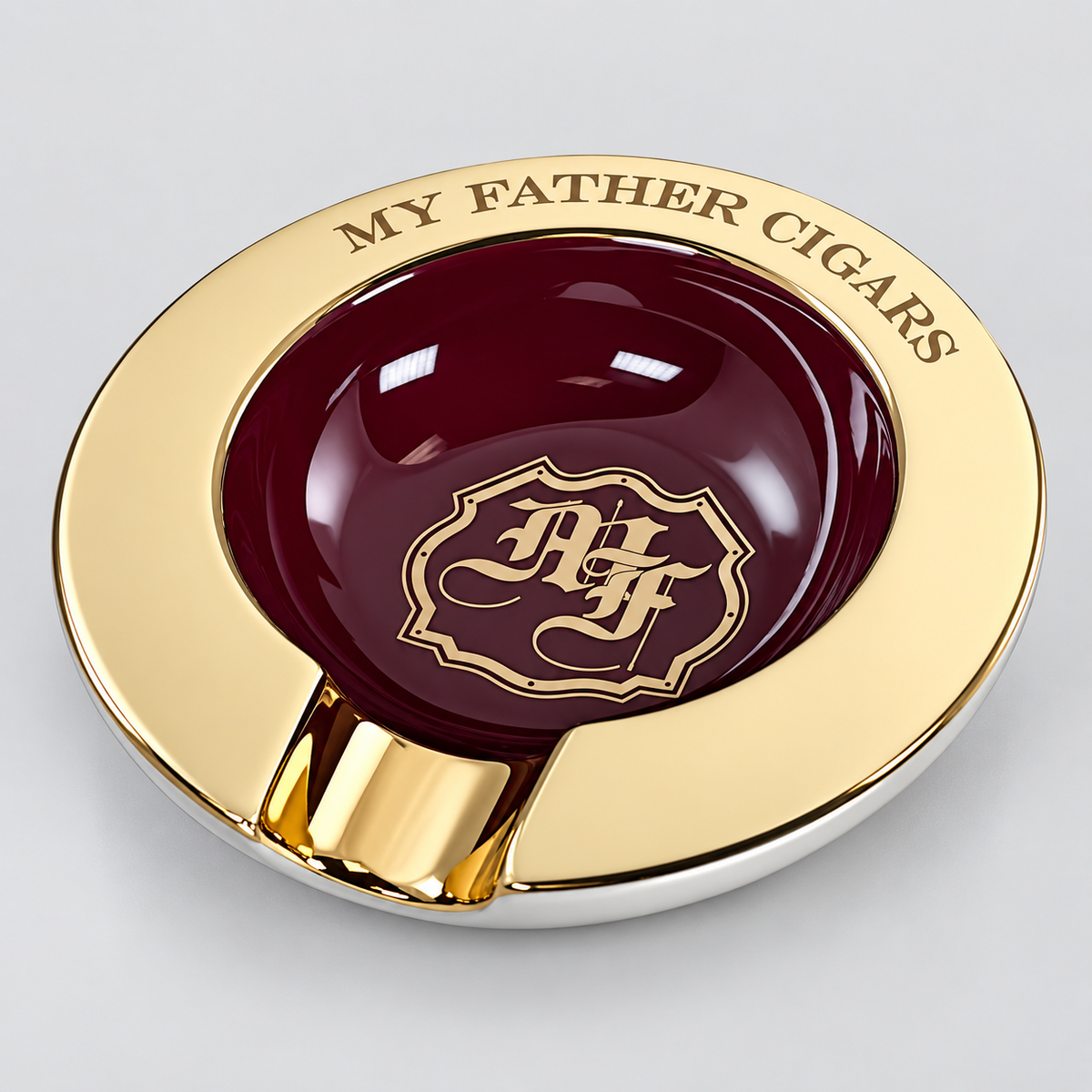 A round gold and silver ashtray with a glossy dark red center, featuring the engraved text 'MY FATHER CIGARS' and a matching monogram emblem in the center.