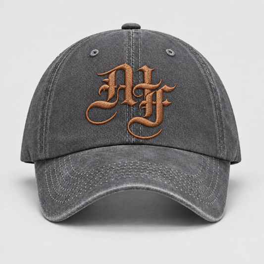 A dark gray washed cotton baseball cap featuring a prominent metallic bronze embroidered monogram logo on the front, set against a neutral light gray background.