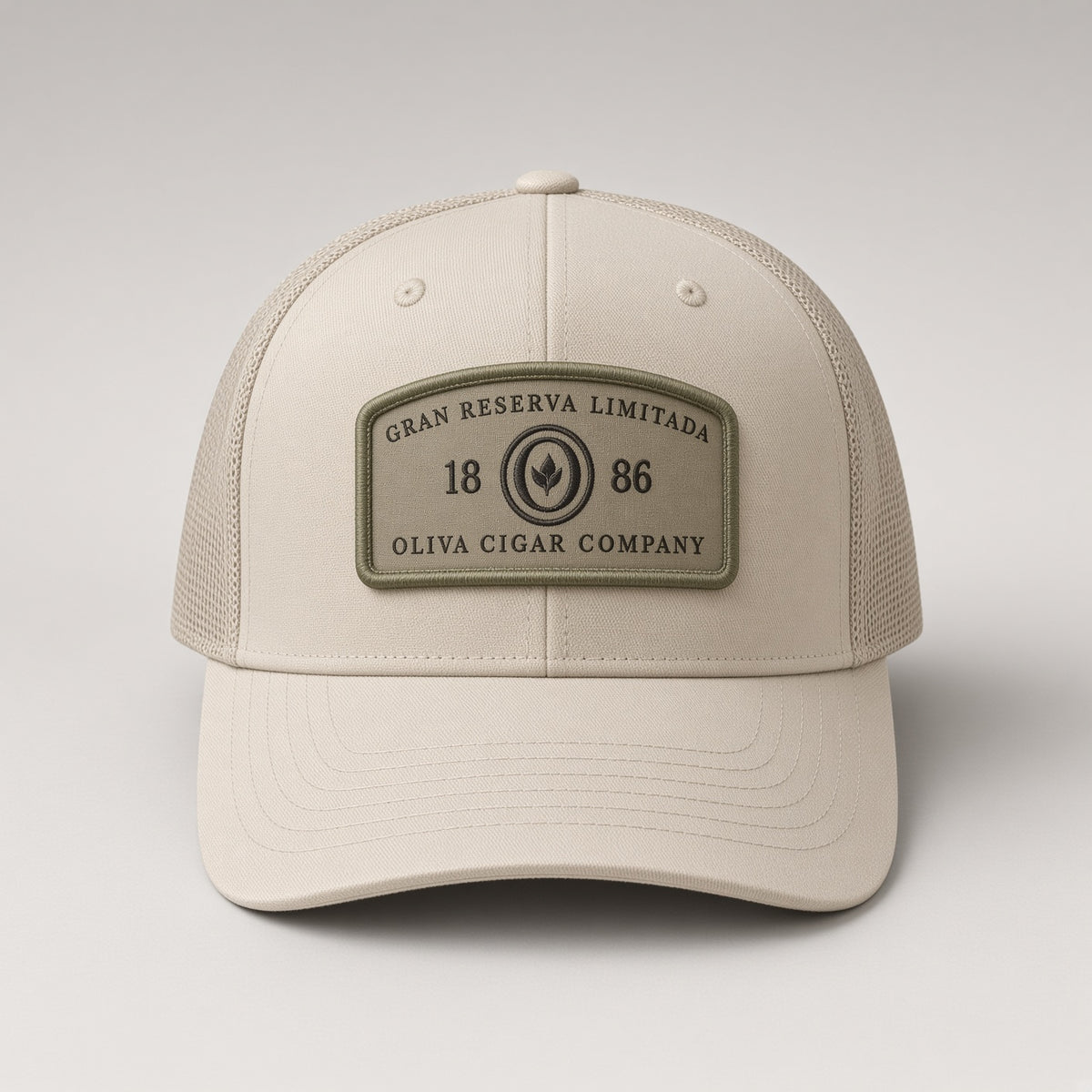 A beige trucker-style baseball cap with a structured front panel and mesh side/back panels. The front features a rectangular olive green patch reading 'GRAN RESERVA LIMITADA 1886 OLIVA CIGAR COMPANY.