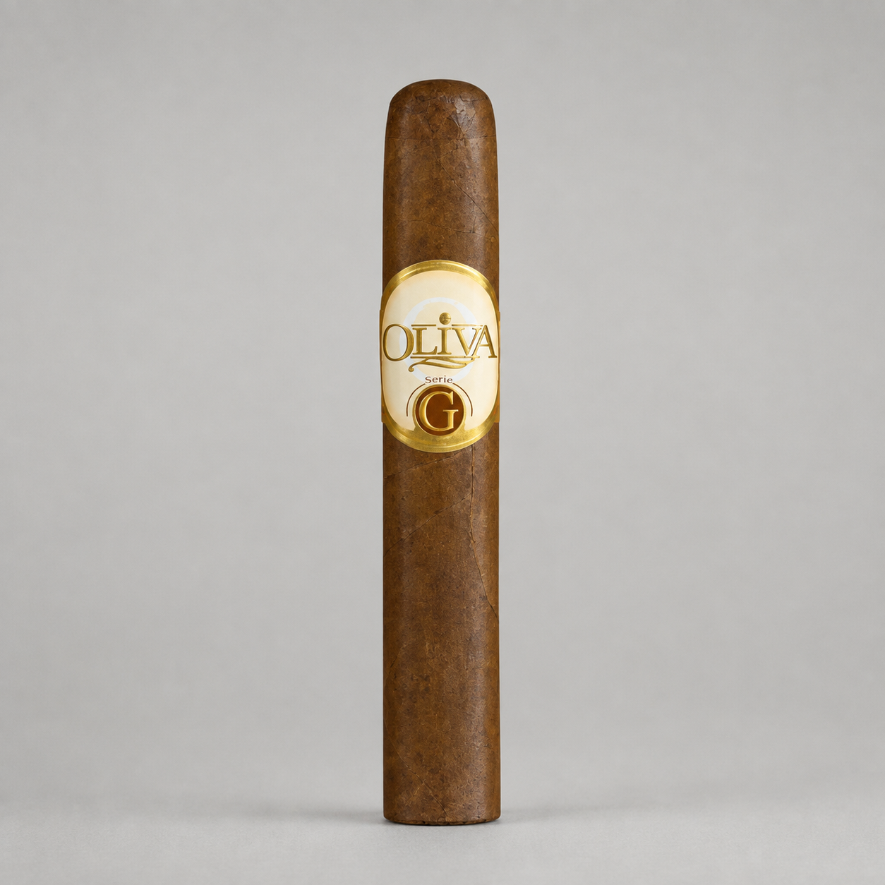 A single brown cigar standing upright against a light gray studio background, featuring a cream and gold oval label with the brand name OLIVA and a large G.