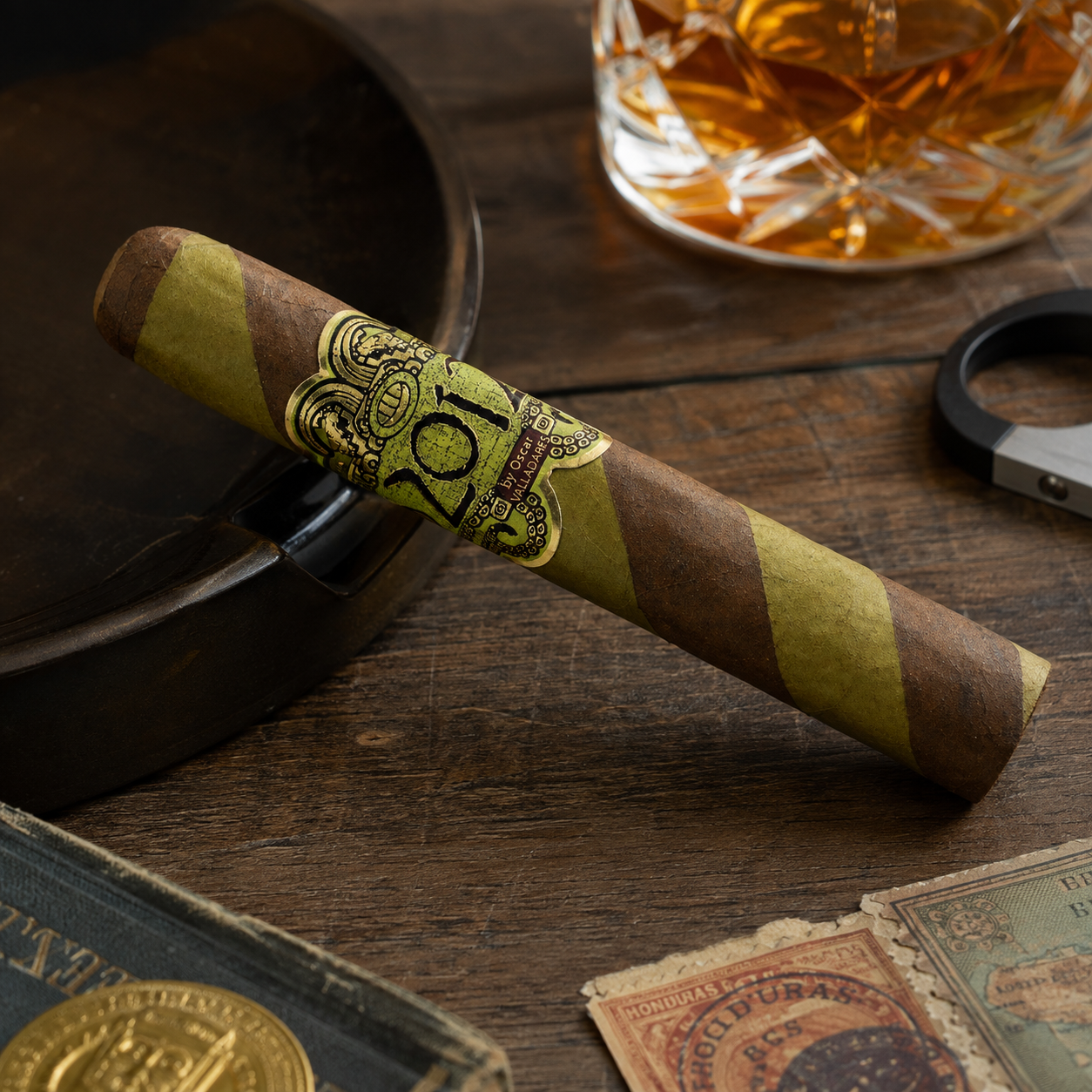 A single cigar resting diagonally on a dark wooden surface, featuring brown and olive-green diagonal stripes with a prominent gold band displaying the number 20. The background includes a glass of amber liquor, a dark ashtray, and vintage stamps.