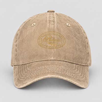 A washed beige baseball cap with a tonal gold embroidered oval logo on the front, photographed against a neutral light gray background.