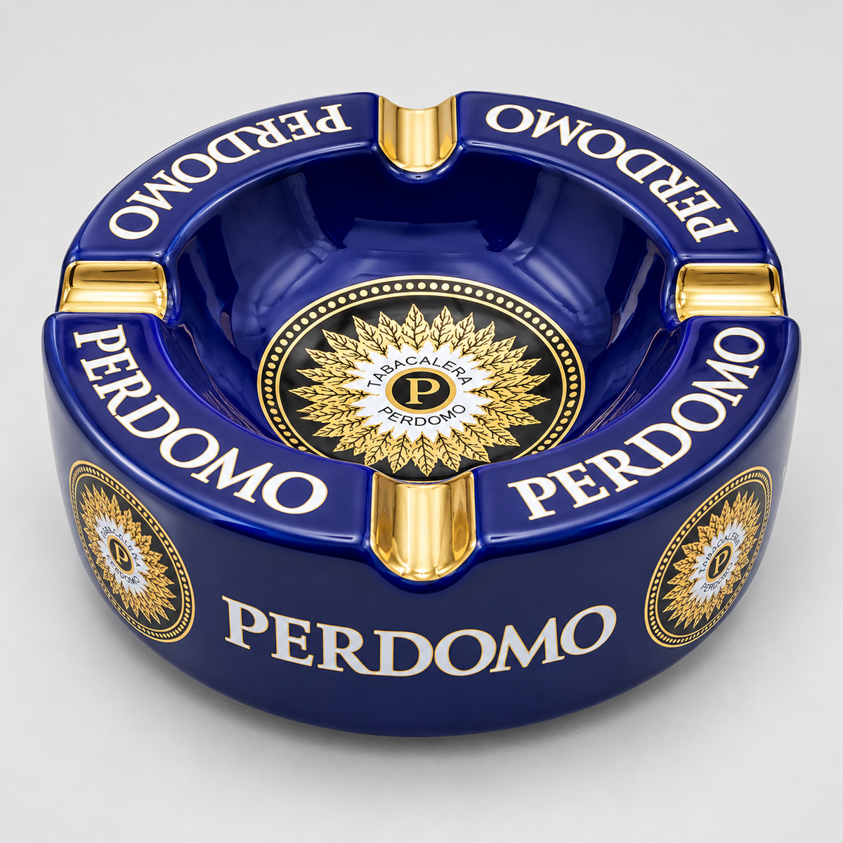 A glossy dark blue ceramic ashtray with gold and white details, featuring the brand name 'PERDOMO' printed repeatedly around the rim and a circular gold emblem in the center.
