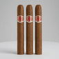 Three brown cigars standing upright side by side on a light gray surface against a light gray background. Each cigar has a red and white band with the text 'MILANO PREMIUM CIGARS' and 'Santa Fe, N.
