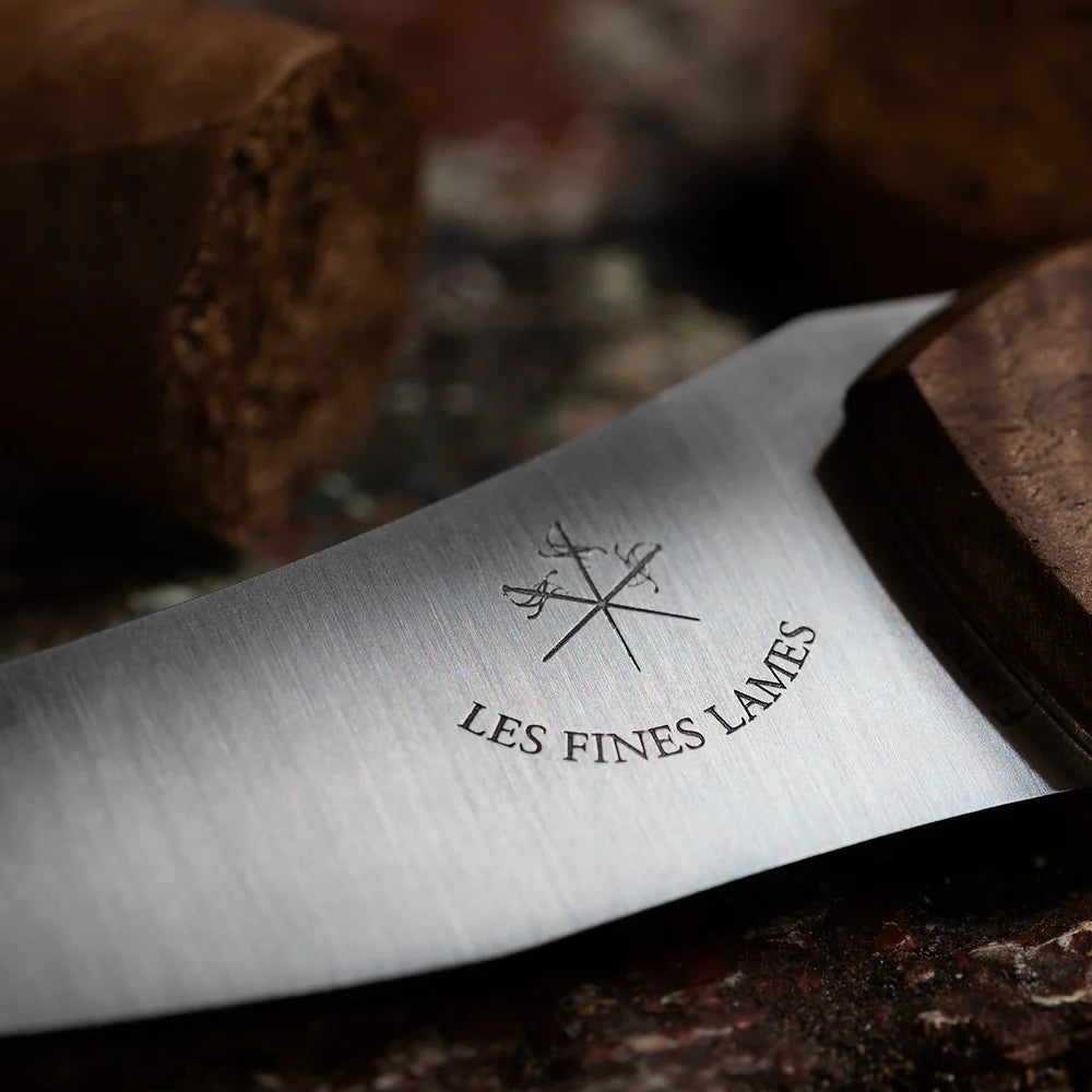 Close-up of a silver metal knife blade engraved with the logo and text 'LES FINES LAMES' and crossed knives, resting on a dark textured surface with blurred brown objects in the background.
