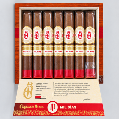 An open wooden cigar box containing eight cigars, with the left cigar removed to show the inner packaging and printed insert. The cigars have brown wrappers with cream and red labels reading 'CRONOS MIL DIAS.