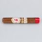 A single cigar in clear plastic wrapping, featuring a white and gold band with red details and a red ribbon tip, photographed horizontally against a solid light gray background.