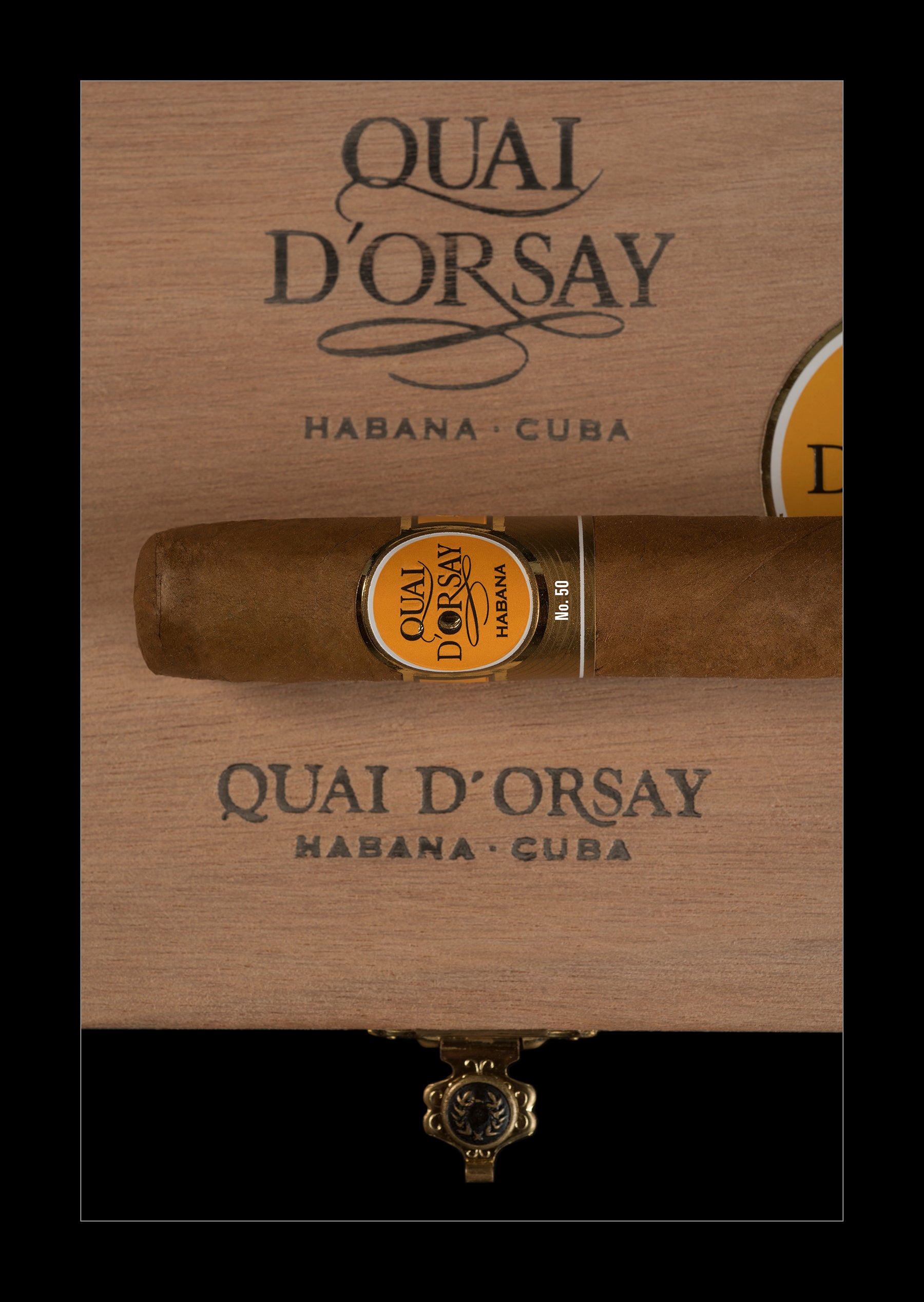 A single cigar resting horizontally on a branded wooden box lid, with the Quai D'Orsay Habana - Cuba branding visible above and below. The cigar has a gold and black band label, and the box is shown against a black background.
