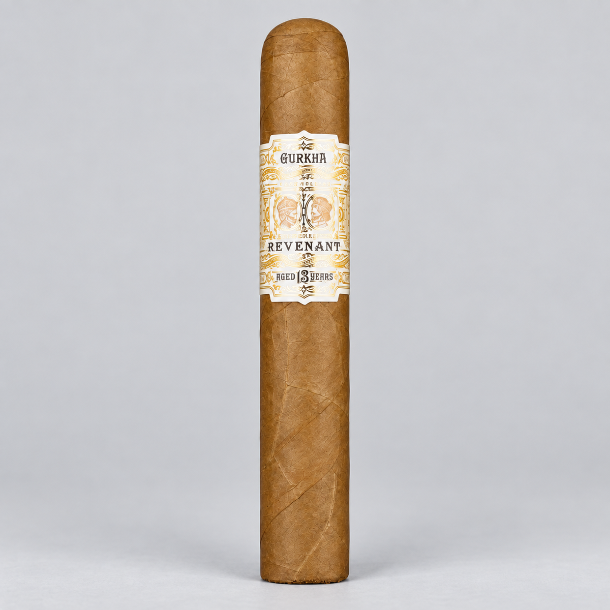 A single Gurkha Revenant cigar standing upright against a light gray studio background. The cigar has a natural brown wrapper with a cream and gold paper band featuring the brand name and illustration.