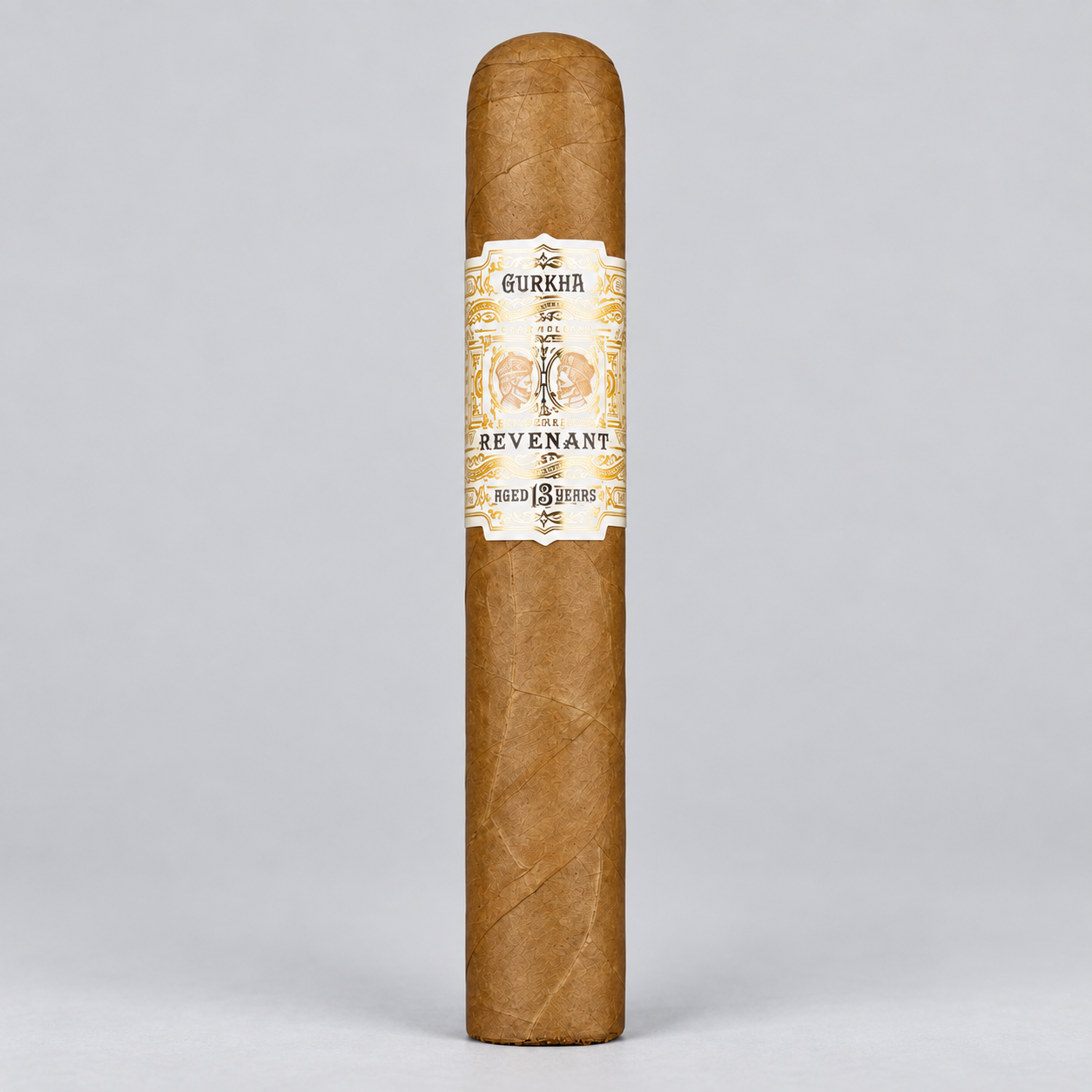 A single Gurkha Revenant cigar standing upright against a light gray studio background. The cigar has a natural brown wrapper with a cream and gold paper band featuring the brand name and illustration.