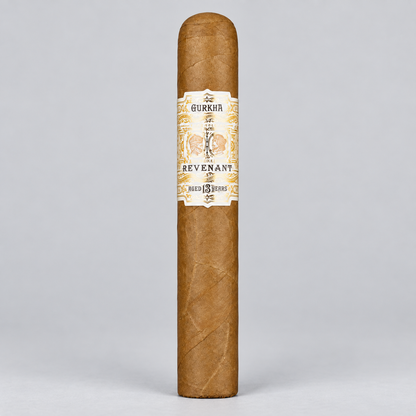 A single Gurkha Revenant cigar standing upright against a light gray studio background. The cigar has a natural brown wrapper with a cream and gold paper band featuring the brand name and illustration.