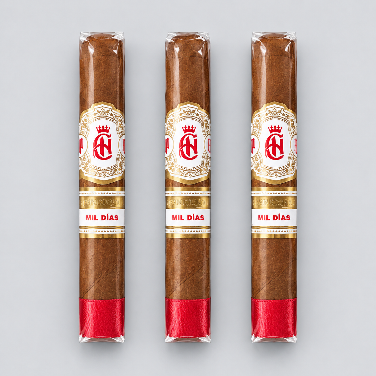 Three cigars in clear plastic wrappers with white and gold labels reading 'MIL DÍAS' and a red band at the base, arranged vertically on a light gray background.