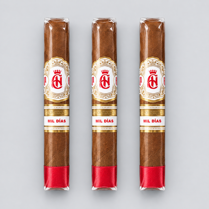 Three cigars in clear plastic wrappers with white and gold labels reading 'MIL DÍAS' and a red band at the base, arranged vertically on a light gray background.
