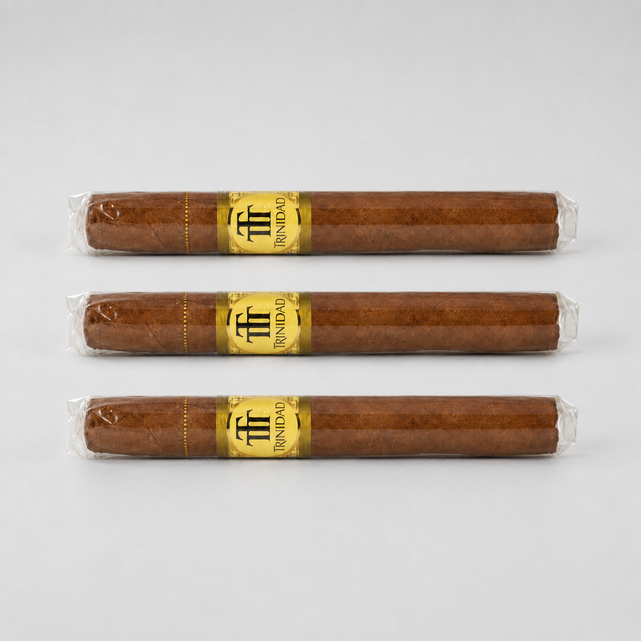 Three brown cigars in clear plastic wrapping with gold and black bands, arranged horizontally on a light gray background.