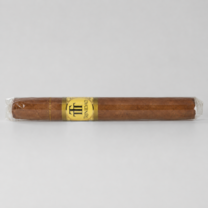 A single brown cigar in clear plastic wrapping with a gold and black Triolo band, photographed horizontally against a light gray background.