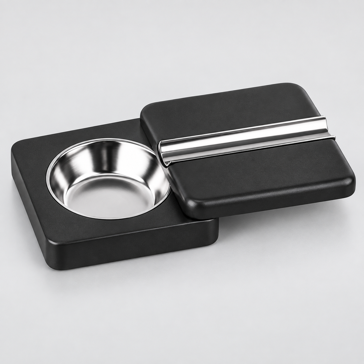 A black rectangular cigar holder with a circular stainless steel bowl on one side and a matching lid on top featuring a silver metal accent bar, shown on a light gray background.