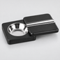 A black rectangular cigar holder with a circular stainless steel bowl on one side and a matching lid on top featuring a silver metal accent bar, shown on a light gray background.