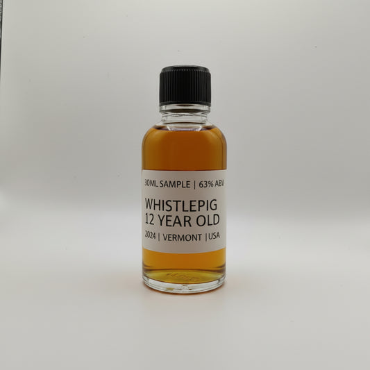 Small bottle of WhistlePig 12 Year Old whiskey sample on a white surface with a gray leather chair in the background.