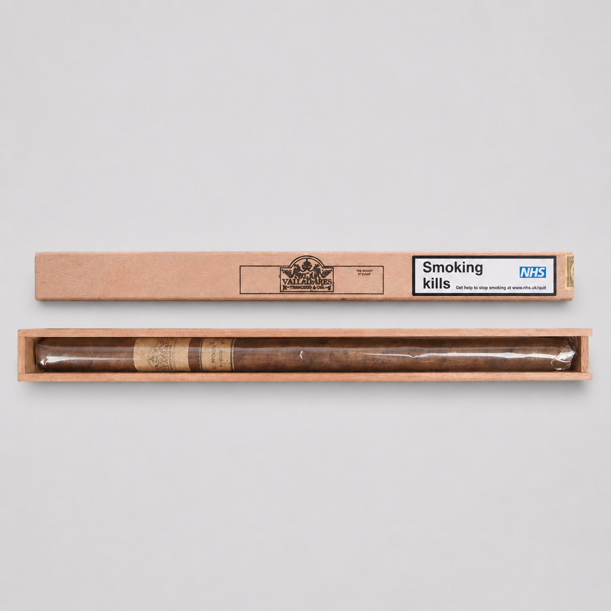 A long cigar in clear plastic wrapping with brown paper bands, displayed horizontally in an open light wood box. The box lid is placed above, showing a printed label that reads 'Smoking kills' alongside the NHS logo and a circular brand emblem.