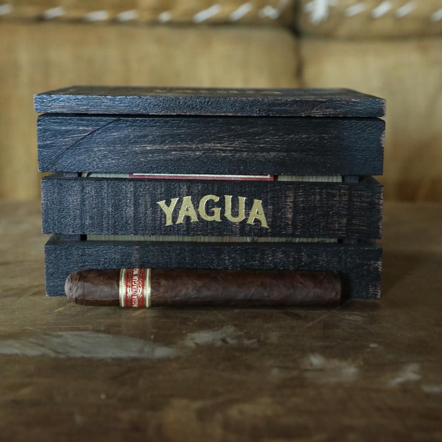 A dark wooden cigar box with the brand name YAGUA in gold lettering, with a dark brown cigar resting horizontally in front of it on a rustic wooden surface.