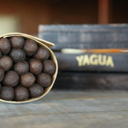 A small paper-wrapped bundle of dark brown cigar tips sits on a rustic wooden surface, with a blurred stack of cigar boxes in the background, including one labeled YAGUA.