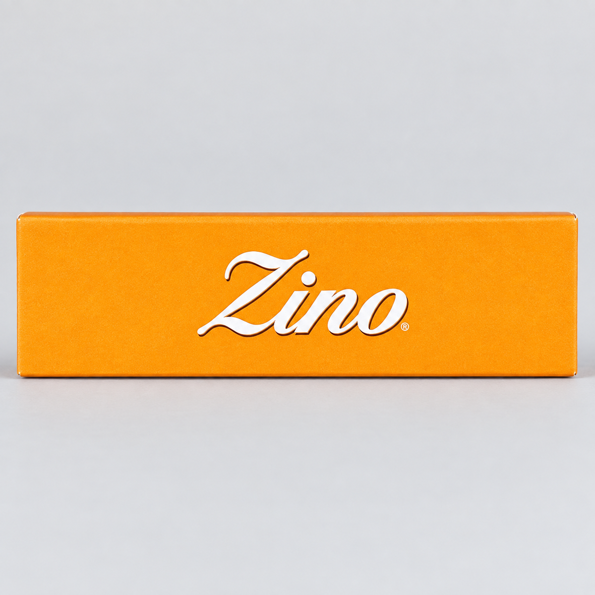 A bright orange rectangular box with the brand name 'Zino' in white cursive script, photographed against a light gray background.