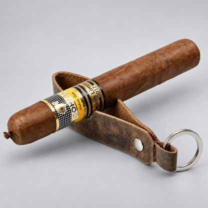 A brown cigar resting on a brown leather key holder with a silver key ring, set against a light gray background. The cigar features a gold and black band labeled 'Cigars de lujo' by 'A.