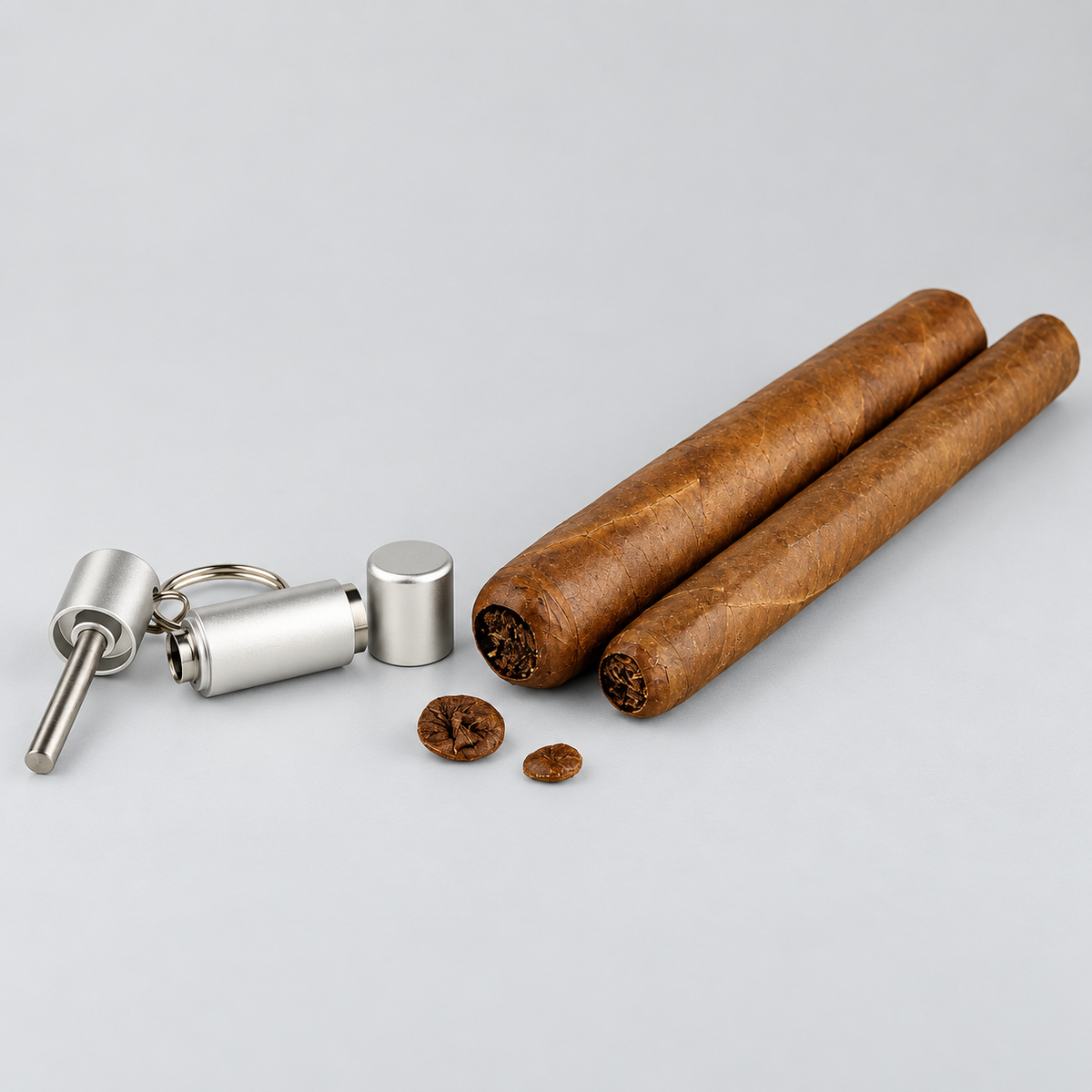 Two brown cigars and a silver metal cigar cutter keychain set on a light gray background.
