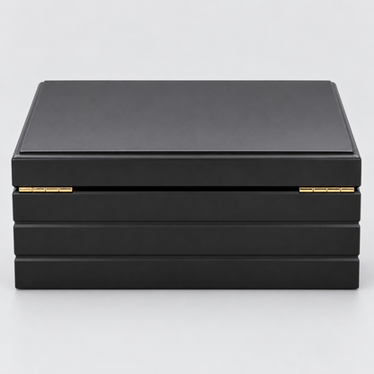 A black rectangular HUMIDOR box with a hinged lid and small gold-tone metal accents on the front, photographed on a clean light gray background.