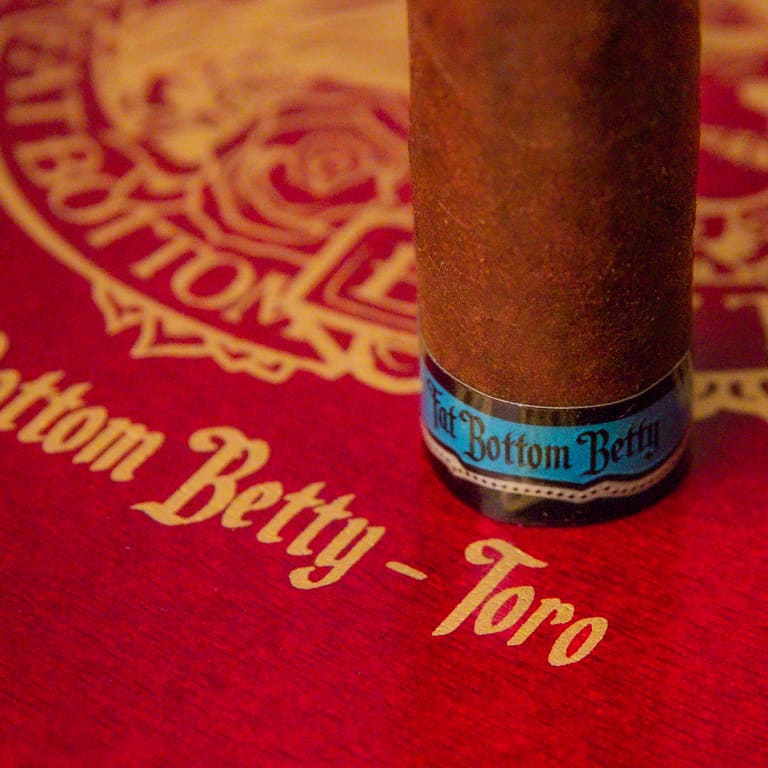 A close-up of a brown cigar standing upright on a red textured surface with gold script reading 'Fat Bottom Betty - Toro' and a matching circular emblem in the background. The cigar has a blue band with black lettering that reads 'Fat Bottom Betty'.