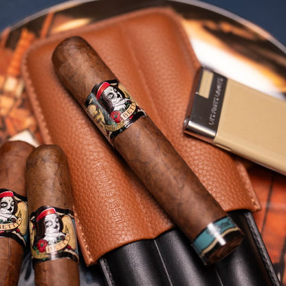 A close-up of premium cigars in a brown leather pouch, with a gold and black lighter nearby. The cigars feature distinctive black, red, and gold bands with a skull illustration and the brand name 'Les Fines'.