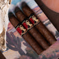Three dark brown cigars with red and black illustrated bands featuring a skeleton woman, arranged on a colorful printed fabric surface.