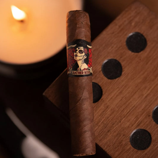 A single brown cigar with a decorative paper label reading 'LEATHER ROSE' and showing a stylized face illustration, standing upright on a wooden surface with blurred warm lighting in the background.
