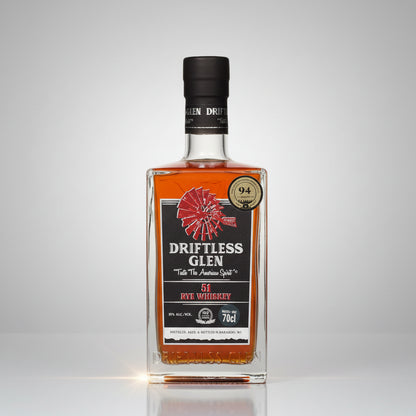 A bottle of Driftless Glen51 Rye Whiskey on a white background with a soft reflection. The bottle is clear glass showing the amber liquid inside, with a black cap and a label featuring orange, black, and red graphics.