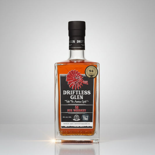 A bottle of Driftless Glen51 Rye Whiskey on a white background with a soft reflection. The bottle is clear glass showing the amber liquid inside, with a black cap and a label featuring orange, black, and red graphics.