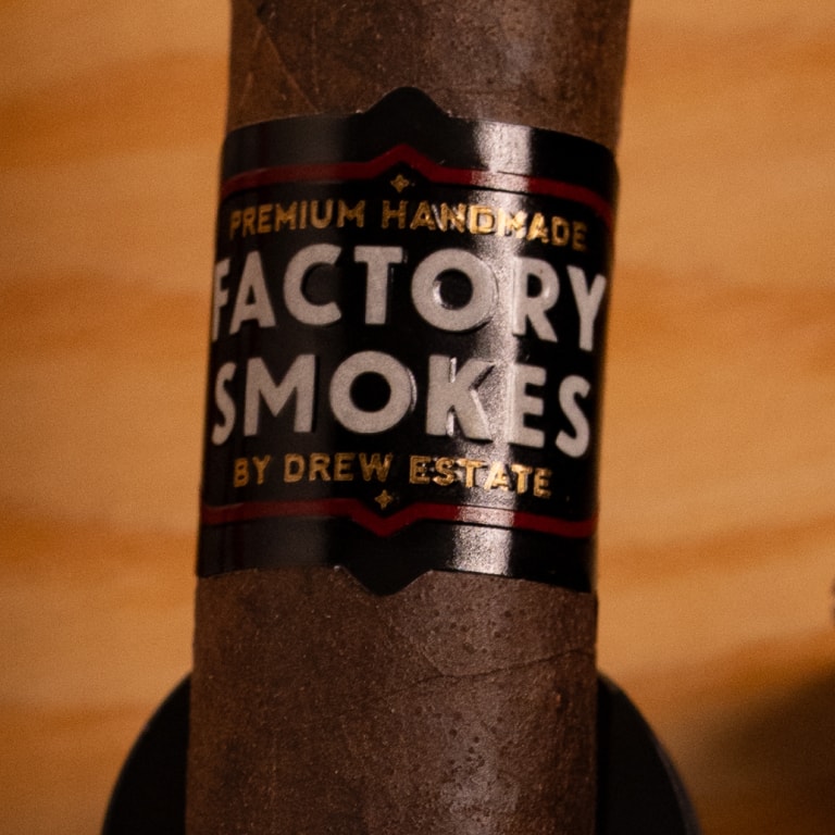 A close-up of a dark brown cigar with a black paper band reading 'Factory Smokes' and 'By Drew Estate' on a warm wooden background.