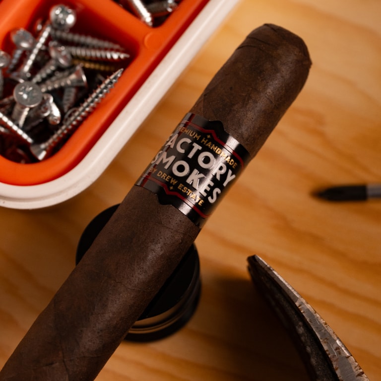 A dark brown cigar with a black and white paper band reading 'Factory Smokes' is shown on a wooden surface, with a small orange-and-white container of screws in the background and a cigar cutter in the foreground.