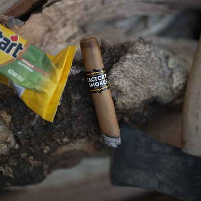 A single cigar with a black and gold 'Factory Smokes' band rests on a piece of driftwood next to a yellow and green package, with a dark axe handle in the blurred background.