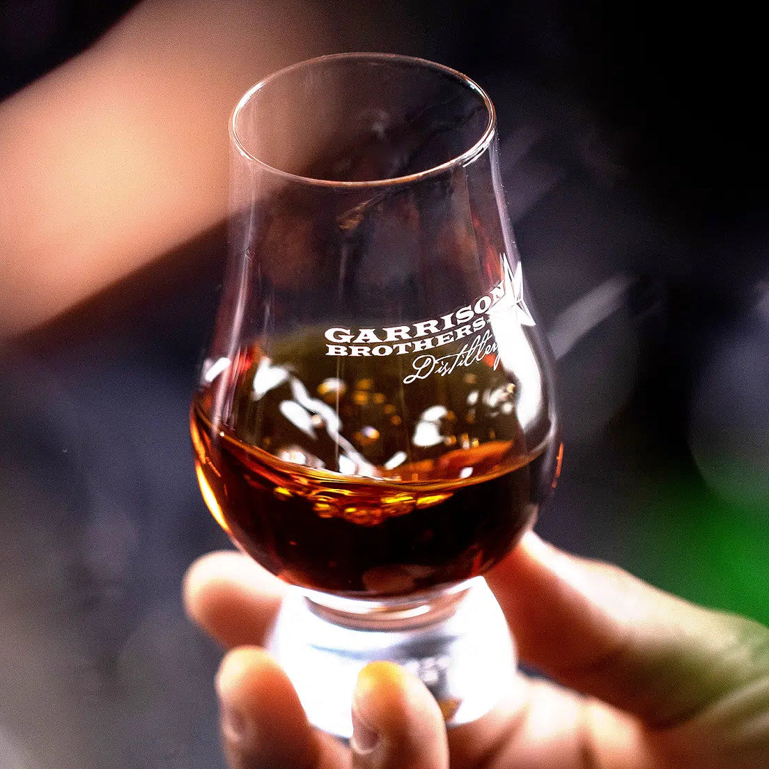 Hand holding a glass of whiskey with 'Garrison Brothers' branding against a blurred background