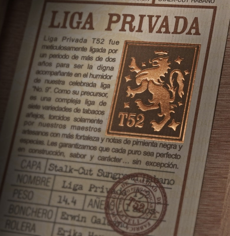 A close-up of a Liga Privada T52 cigar packaging card, showing the brand name, Spanish descriptive text, a gold-toned crest with 'T52', and a circular stamp on a light wood background.