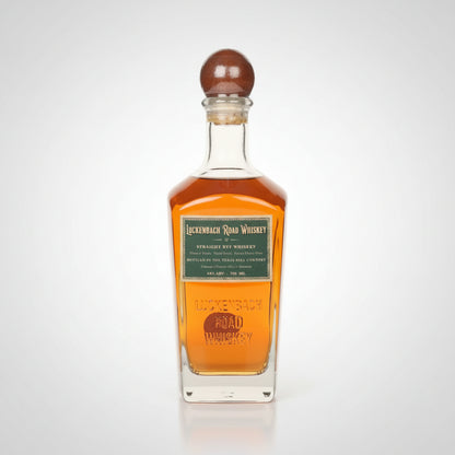 A bottle of Luckenbach Rye Road Whiskey with amber liquid, a green label, and a brown wooden stopper, shown on a white background.