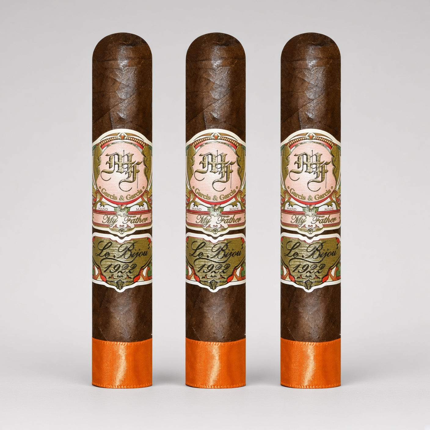 Three dark brown cigars standing upright side by side on a light gray background. Each cigar has a decorative cream, green, and red band label reading 'DH' and 'La Royale 1925', with a bright orange band at the bottom.