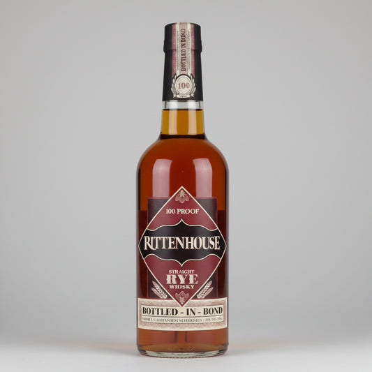 A bottle of Rittenhouse Straight Rye Whiskey on a white background with a soft reflection. The amber liquid inside the clear glass bottle is visible, along with a dark brown and beige label and a gold neck seal.