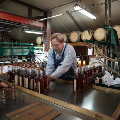 A man in an apron works at a bottling line inside a brewery or distillery, handling rows of clear glass bottles filled with amber liquid. Wooden barrels and industrial equipment are visible in the background.